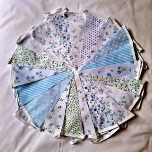 May include: A fabric bunting banner featuring a collection of triangular pennants in various shades of blue and white. The patterns include floral prints, polka dots, and thin stripes, all attached to a white fabric header tape.