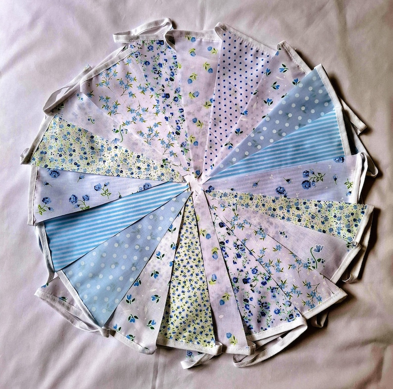 May include: A fabric bunting banner featuring a collection of triangular pennants in various shades of blue and white. The patterns include floral prints, polka dots, and thin stripes, all attached to a white fabric header tape.