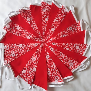 May include: A decorative bunting banner featuring alternating triangular flags in solid red and red with a white floral vine pattern. Each flag is edged with white fabric trim, and the entire set is connected by a white fabric header tape.