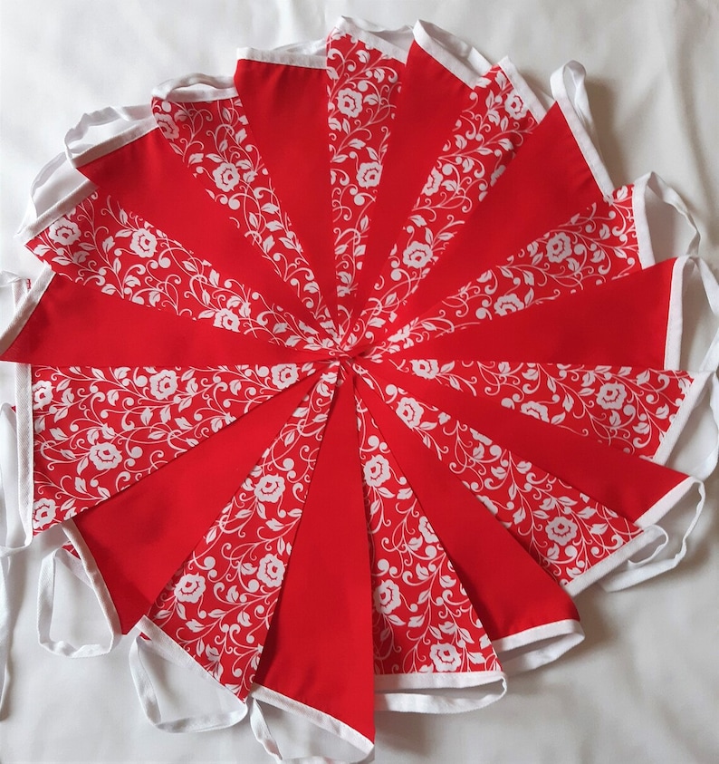 May include: A decorative bunting banner featuring alternating triangular flags in solid red and red with a white floral vine pattern. Each flag is edged with white fabric trim, and the entire set is connected by a white fabric header tape.