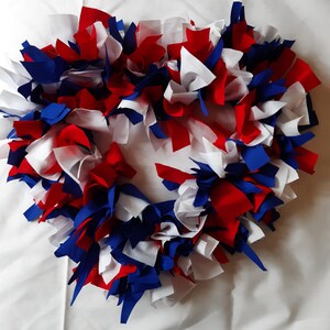 May include: A patriotic wreath made of fabric strips in red, white, and blue. The wreath is circular and has a fluffy, textured appearance, perfect for adding a touch of Americana to any home decor. The colors are vibrant and the design is eye-catching.
