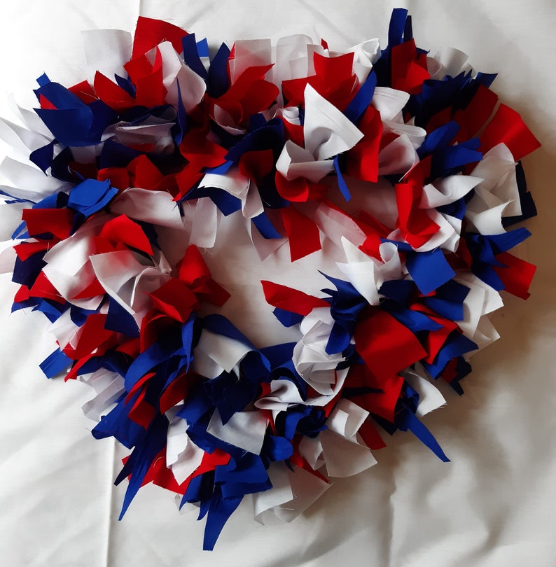 May include: A patriotic wreath made of fabric strips in red, white, and blue. The wreath is circular and has a fluffy, textured appearance, perfect for adding a touch of Americana to any home decor. The colors are vibrant and the design is eye-catching.