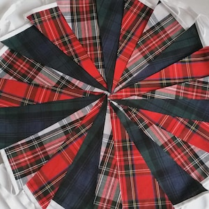 May include: A circular arrangement of triangular fabric pennants featuring various red, green, and blue plaid tartan patterns. Each pennant is edged with white fabric trim, creating a decorative bunting display with multiple intersecting textile designs.