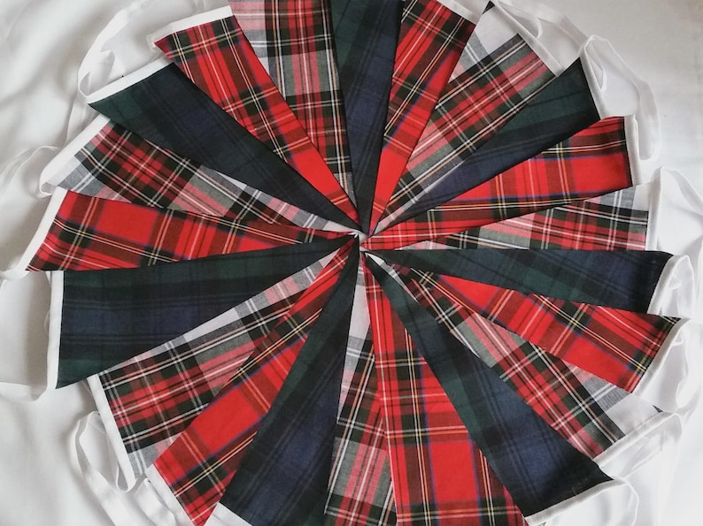 May include: A circular arrangement of triangular fabric pennants featuring various red, green, and blue plaid tartan patterns. Each pennant is edged with white fabric trim, creating a decorative bunting display with multiple intersecting textile designs.