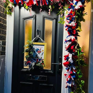 May include: A black front door decorated with red, white, and blue fabric streamers and greenery. A framed picture with the text "Kings Coronation" and a Union Jack flag is hanging on the door. The door has gold-toned window panels.