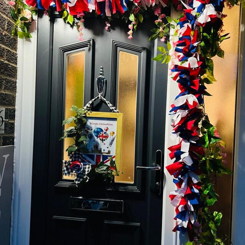 May include: A black front door decorated with red, white, and blue fabric streamers and greenery. A framed picture with the text "Kings Coronation" and a Union Jack flag is hanging on the door. The door has gold-toned window panels.