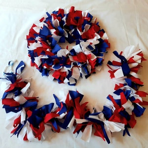 May include: A patriotic wreath and garland set. The wreath is circular, and the garland is long and flowing. Both are made of red, white, and blue fabric strips, perfect for Independence Day or other patriotic holidays.