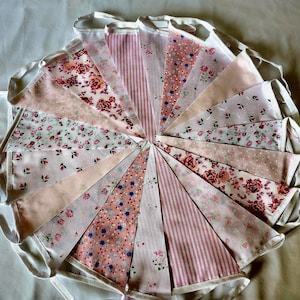 May include: A circular arrangement of fabric bunting flags featuring various patterns in shades of pink, white, and red. The triangular pennants display floral prints, stripes, and polka dots, all connected by a white fabric tape border.