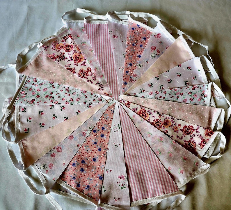 May include: A circular arrangement of fabric bunting flags featuring various patterns in shades of pink, white, and red. The triangular pennants display floral prints, stripes, and polka dots, all connected by a white fabric tape border.