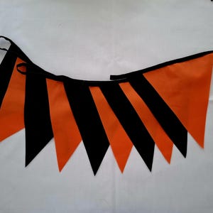 May include: A festive orange and black pennant banner. The triangular flags alternate between solid orange and black stripes, strung together with a black cord. Perfect for Halloween or other celebrations.