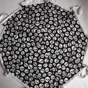 May include: Black apron with a white skull and crossbones pattern. The apron has white trim and ties. The design features numerous white skulls on a black background, creating a spooky aesthetic.