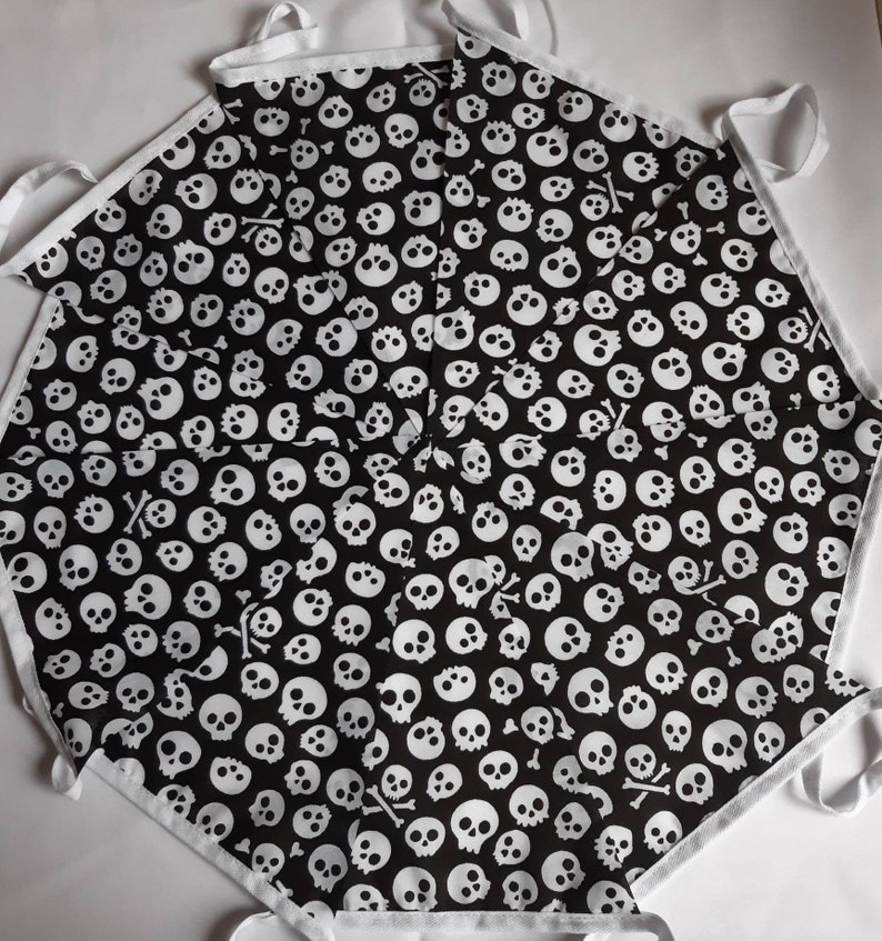 May include: Black apron with a white skull and crossbones pattern. The apron has white trim and ties. The design features numerous white skulls on a black background, creating a spooky aesthetic.