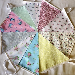 May include: A fabric bunting banner featuring ten triangular flags arranged in a circular pattern. Each flag displays a different floral print in various colors including pink, light blue, yellow, green, white, and grey, all attached to a white tape.