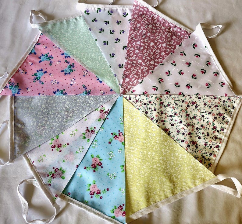 May include: A fabric bunting banner featuring ten triangular flags arranged in a circular pattern. Each flag displays a different floral print in various colors including pink, light blue, yellow, green, white, and grey, all attached to a white tape.