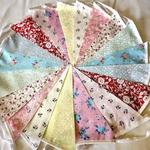 May include: A bunting banner featuring a collection of triangular fabric flags arranged in a circular pattern. The flags display various floral patterns in colours including red, pink, yellow, blue, green, and white, all attached to a white ribbon.