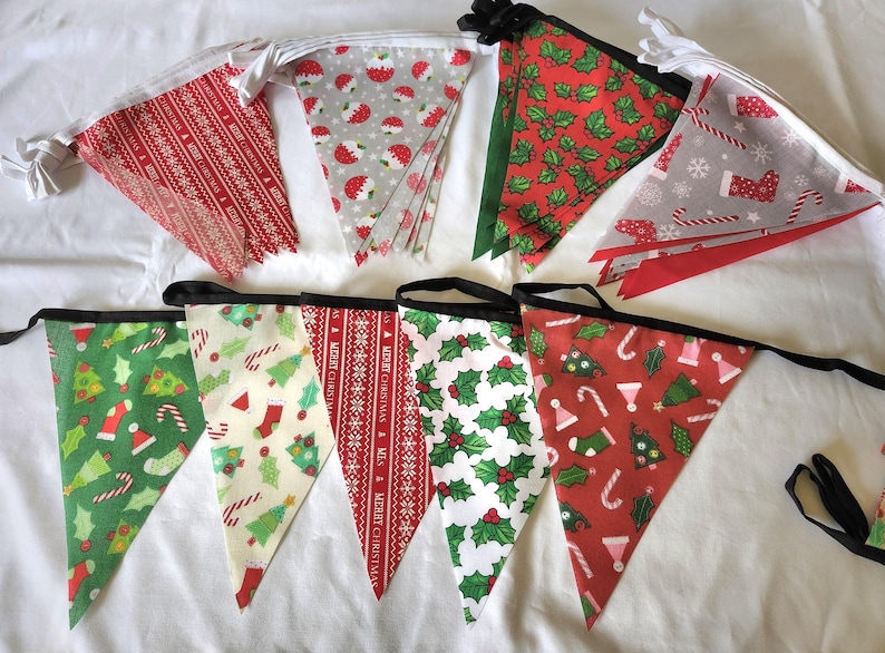 May include: A set of triangular fabric bunting flags featuring various Christmas-themed patterns including red and white Merry Christmas text, green holly leaves, candy canes, stockings, and Christmas trees, all attached to a black or white ribbon.