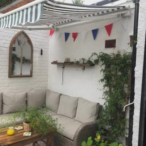 May include: Outdoor patio setting with a striped awning, a wooden table, and a wicker sofa with beige cushions. A wooden shelf displays small decorative items. Red, white, and blue bunting hangs on the wall. A large mirror is on the left.