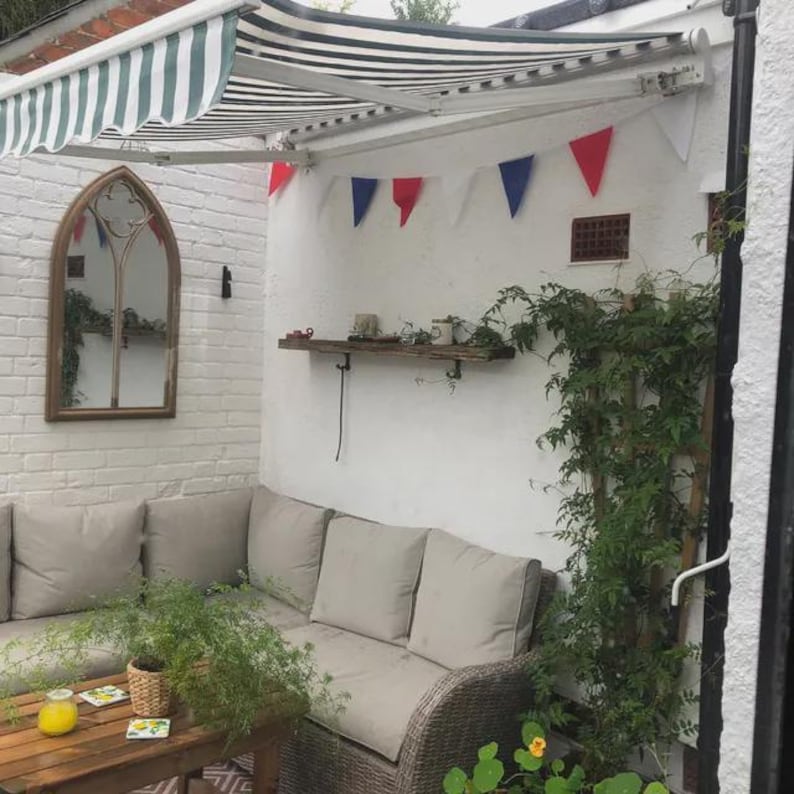 May include: Outdoor patio setting with a striped awning, a wooden table, and a wicker sofa with beige cushions. A wooden shelf displays small decorative items. Red, white, and blue bunting hangs on the wall. A large mirror is on the left.