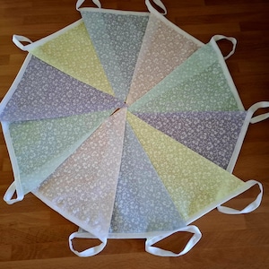 May include: A fabric bunting banner featuring a series of triangular pennants in pastel shades of yellow, blue, pink, and green. Each pennant displays a delicate white floral vine pattern and is bordered by a white fabric trim with loops.
