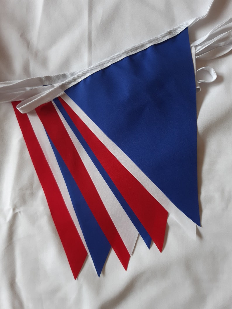 May include: A string of triangular bunting flags in red, white, and blue. The flags are made of fabric and are attached to a white ribbon. The bunting is likely for a patriotic or celebratory event.