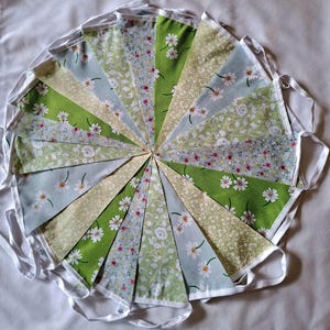 May include: A fabric bunting banner featuring triangular flags arranged in a circular pattern. The flags display various patterns including green polka dots with white daisies, light blue floral prints, and intricate white vine motifs.