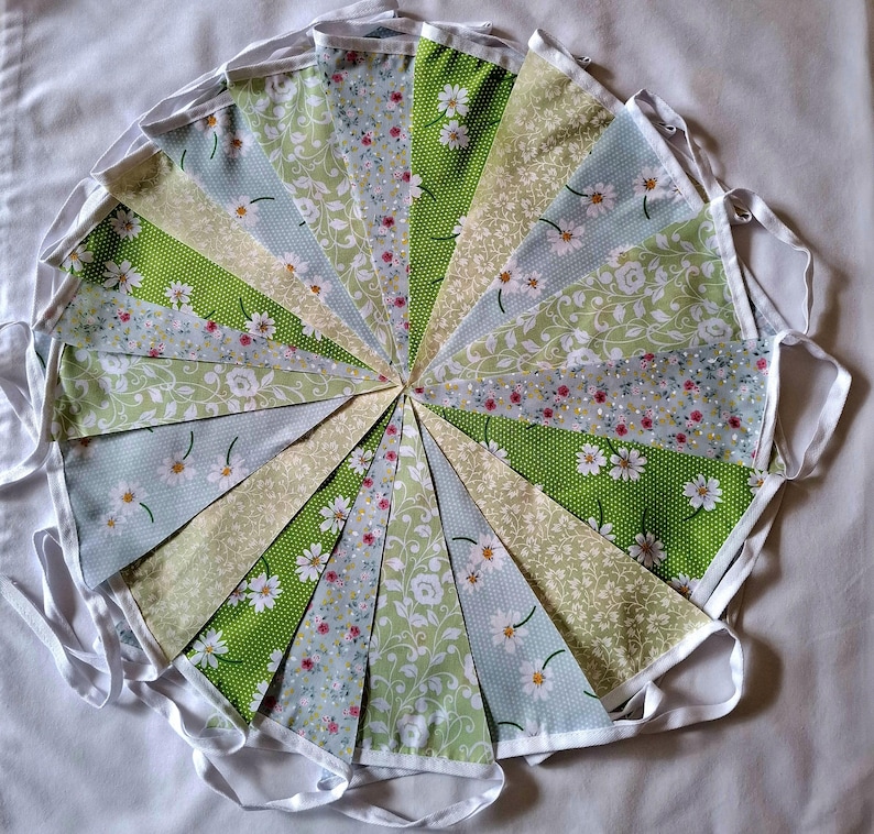 May include: A fabric bunting banner featuring triangular flags arranged in a circular pattern. The flags display various patterns including green polka dots with white daisies, light blue floral prints, and intricate white vine motifs.