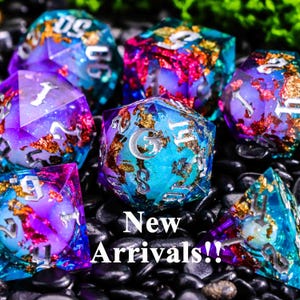 以下が含まれることがあります： 青、紫、金色のカラフルな宝石のようなサイコロのコレクション。サイコロは黒い石の上に散らばっています。「New Arrivals!!」というテキストが白で表示されています。