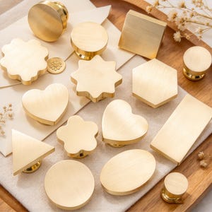 May include: A collection of twelve brass-toned metal wax seal stamps in various geometric and organic shapes, including circles, squares, hexagons, hearts, stars, flowers, triangles, and rectangles, all featuring a smooth, brushed finish.