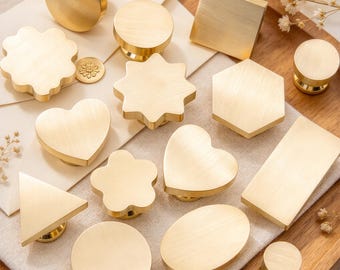 May include: A collection of twelve brass-toned metal wax seal stamps in various geometric and organic shapes, including circles, squares, hexagons, hearts, stars, flowers, triangles, and rectangles, all featuring a smooth, brushed finish.