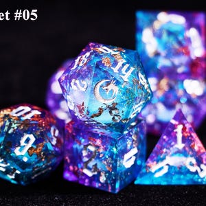 May include: Set of seven polyhedral dice with a celestial theme. The dice are a mix of blue, purple, and gold, with white numbering. The image includes a D20, D12, D10, D8, D6, D4, and a D10 percentile die. The text "Set #05" is in the upper left corner.