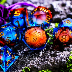 Puede incluir: Un conjunto de dados poliédricos coloridos, perfectos para juegos de mesa. Los dados presentan una vibrante mezcla de tonos azules y naranjas con números dorados. Se encuentran sobre una superficie de piedra gris texturizada.