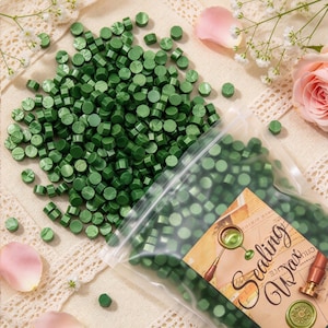 May include: A large collection of small, round, forest green sealing wax beads scattered next to a clear plastic bag filled with more of the same beads. The bag features a label with the text Sealing Wax and an illustration of a wax seal.