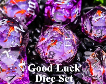 Könnte beinhalten: Ein Satz aus lila und transparenten polyedrischen Würfeln mit orangefarbenen Fischen und silbernen Zahlen. Die Würfel sind vor einem schwarzen Hintergrund mit dem Text "Good Luck Dice Set" platziert.