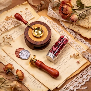 May include: A wax seal kit featuring a wooden handle melting spoon, a circular wooden melting stove, a brass stamp with a wooden handle, and a glass vial filled with small red wax beads. Two finished wax seals are present, one red and one silver.