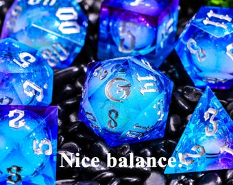 Könnte beinhalten: Eine Sammlung von blauen und violetten Harzwürfeln mit silbernen Zahlen. Die Würfel haben verschiedene Formen, darunter ein D20, D6 und D4. Der Text "Nice balance!" ist sichtbar. Die Würfel liegen auf einem Bett aus schwarzen Steinen.