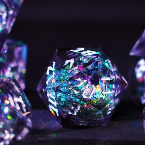Puede incluir: Primer plano de un conjunto de dados poliédricos. Los dados son una mezcla de morado, azul y verde con purpurina iridiscente y numeración blanca. Los dados están diseñados para juegos de rol de mesa.