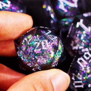 May include: A close-up of a polyhedral die with the number 20 visible. The die is black and filled with colorful glitter. Other dice are visible in the background. The dice are used for tabletop role-playing games.