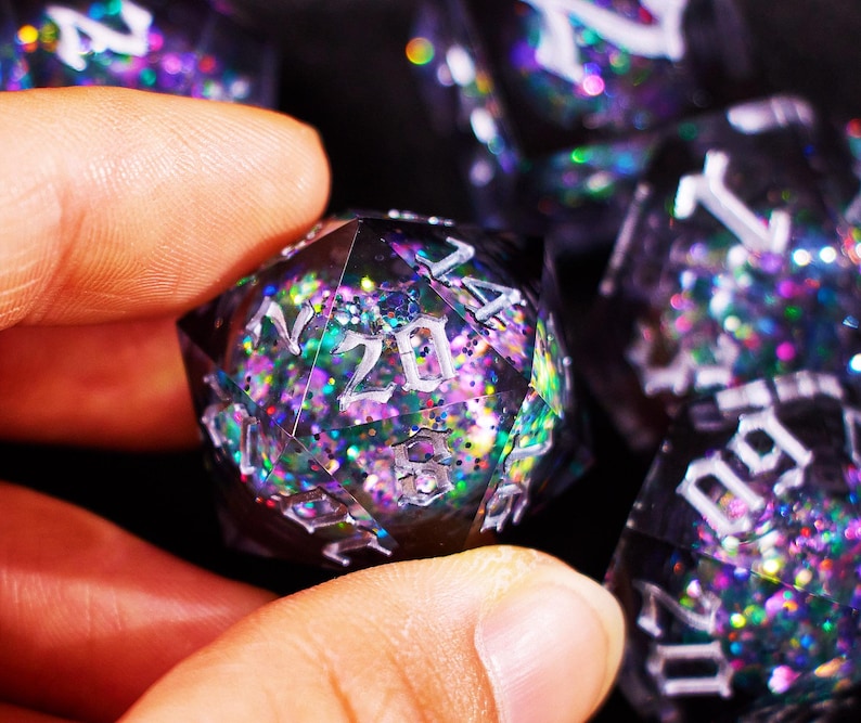 May include: A close-up of a polyhedral die with the number 20 visible. The die is black and filled with colorful glitter. Other dice are visible in the background. The dice are used for tabletop role-playing games.