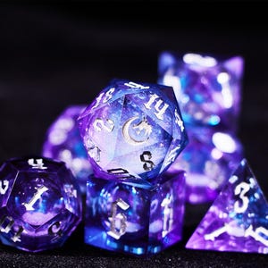 May include: A set of polyhedral dice with a galaxy design. The dice are a mix of purple and blue with silver numbering. The D20 die features a crescent moon and a dragon. The dice are for tabletop games.