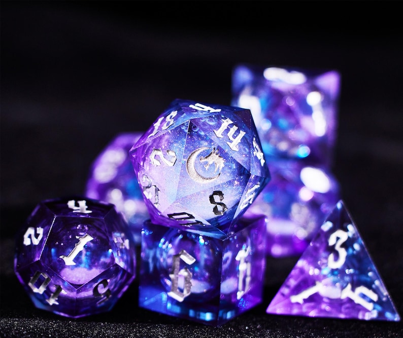 May include: A set of polyhedral dice with a galaxy design. The dice are a mix of purple and blue with silver numbering. The D20 die features a crescent moon and a dragon. The dice are for tabletop games.