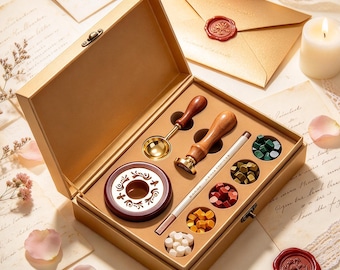 May include: A gold-colored gift box containing a wax seal kit. The set includes a wooden-handled brass stamp, a metal melting spoon, a dark red wax warmer, a white candle, and five small containers filled with wax beads in green, gold, red, yellow, and white.