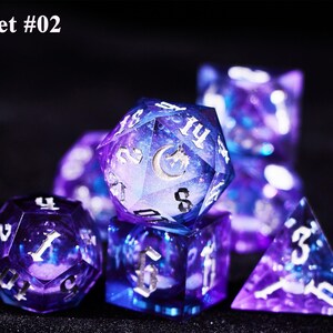May include: Set #02. A set of seven polyhedral dice with a galaxy design in shades of purple and blue. The dice have white numbering and a silver moon and dragon symbol on the largest die.