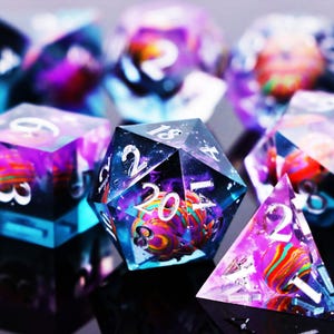 May include: Close-up of several polyhedral dice with a galaxy-like design. The dice are clear with colorful swirls of purple, orange, and blue inside. Each die has white numbers.