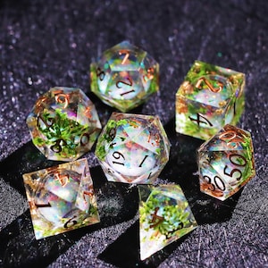 Puede incluir: Juego de siete dados poliédricos para juegos de mesa. Cada dado es transparente con follaje verde incrustado y acentos iridiscentes. Los números son de color cobre. Los dados están dispuestos sobre una superficie oscura y texturizada.