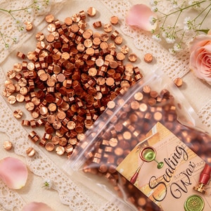 May include: A large collection of small, round, metallic copper-colored wax beads for sealing, with many loose beads scattered and others contained in a clear plastic bag featuring a label with the text Sealing Wax and a graphic illustration.