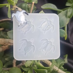 Op de afbeelding: Een witte siliconen mal met vier schildpadvormige holtes en een afgewerkte schildpadhanger. De hanger is zilver en zwart. De mal heeft de letters "ASM" erop gedrukt. De achtergrond bevat groene planten.