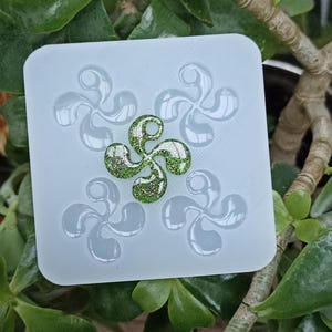 May include: A square, light blue silicone mold with four stylized Basque cross designs. The center cross is filled with green glitter. The mold is set against a backdrop of green leafy plants.