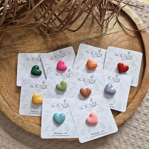 Könnte beinhalten: Eine Sammlung herzförmiger Accessoires in verschiedenen Farben, darunter Grün, Rosa, Orange und Rot, die auf einzelnen Karten präsentiert werden. Die Karten zeigen das Wort "SCHONER" und eine Website-Adresse.