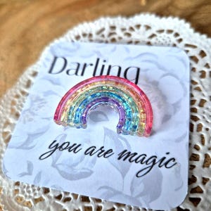 Può includere: Una spilla a forma di arcobaleno con glitter, appoggiata su una carta con la scritta "Darling you are magic". La spilla presenta uno spettro di colori: rosa, giallo, verde, blu e viola. La carta ha un motivo floreale.