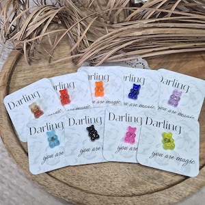 Peut inclure: Collection de breloques en forme d'oursons en gélatine colorés sur de petites cartes. Chaque carte porte le mot "Darling" et la phrase "you are magic". Les oursons sont de différentes couleurs, dont le rouge, l'orange, le bleu et le rose.